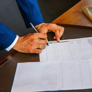 Imagem das mãos de um homem de terno sobre uma mesa de madeira, com caneta em punho, assinando um documento