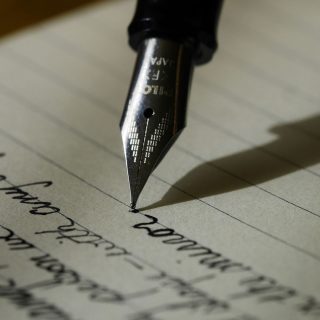 Imagem da ponta de uma caneta de pena escrevendo sobre a folha de um caderno