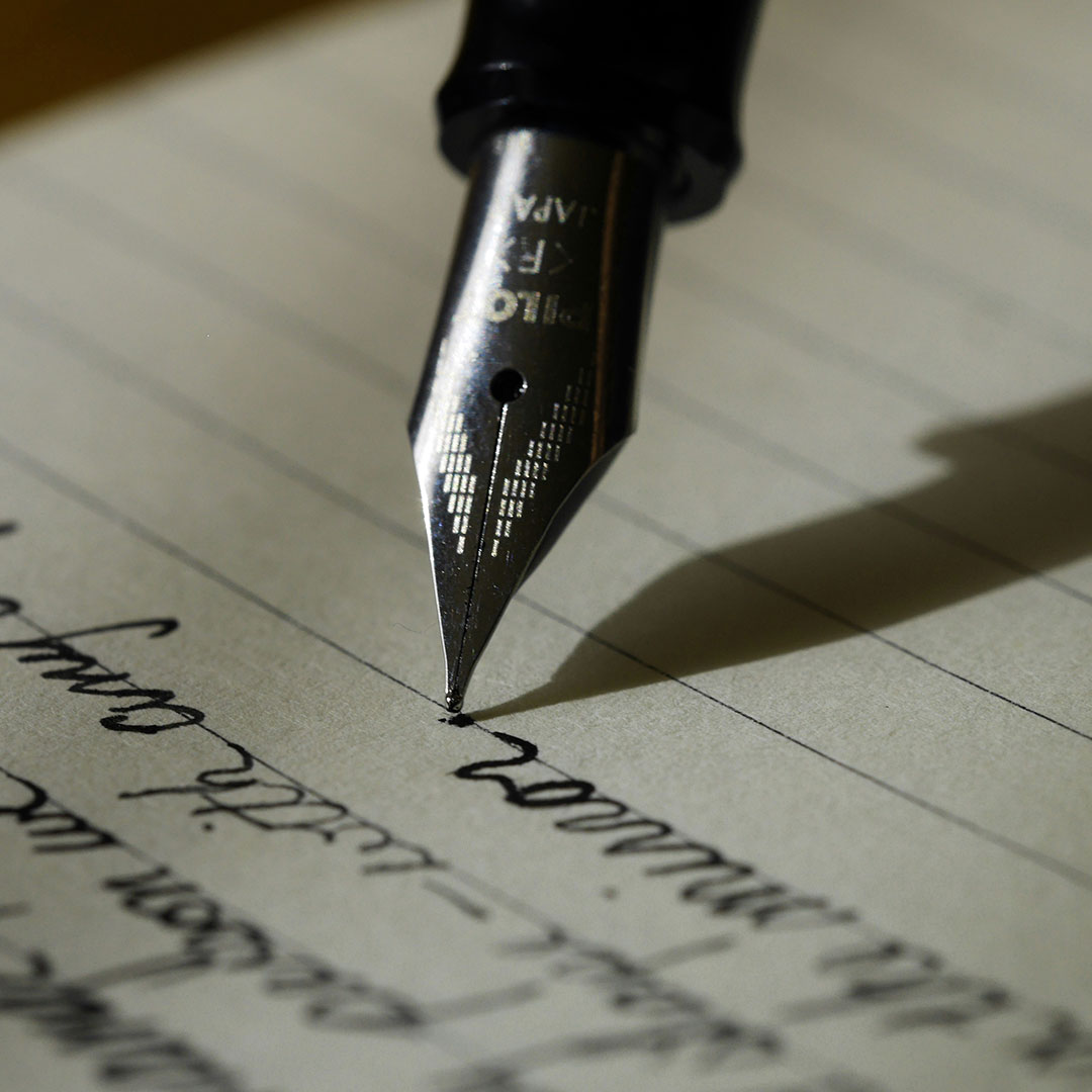 Imagem da ponta de uma caneta de pena escrevendo sobre a folha de um caderno