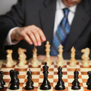 Foto do torso de um homem de terno e gravata movimentando uma peça no tabuleiro de xadrez em sua frente
