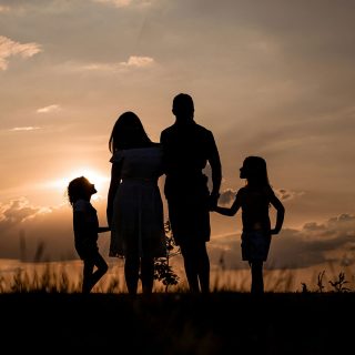 Foto da silhueta de uma família de mãos dadas - composta por um casal com dois filhos - contra a luz do sol